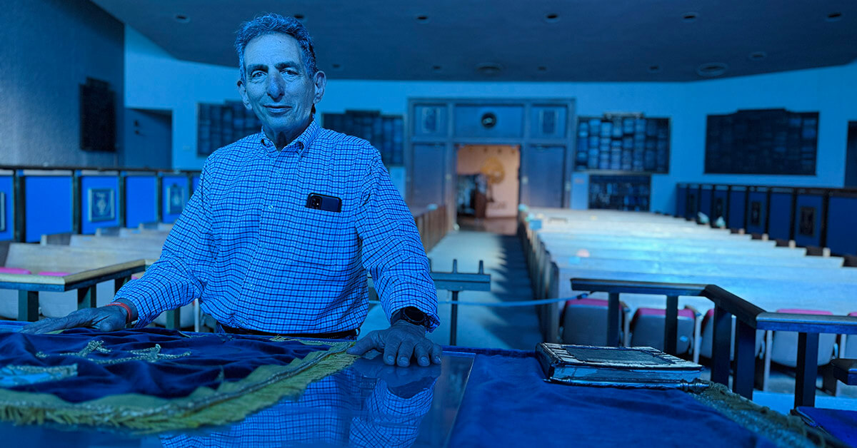 Alan Iszauk at the bimah where he was bar mitzvahed, married, and still prays. The sanctuary's blue stained-glass windows cast an ethereal glow onto the room.