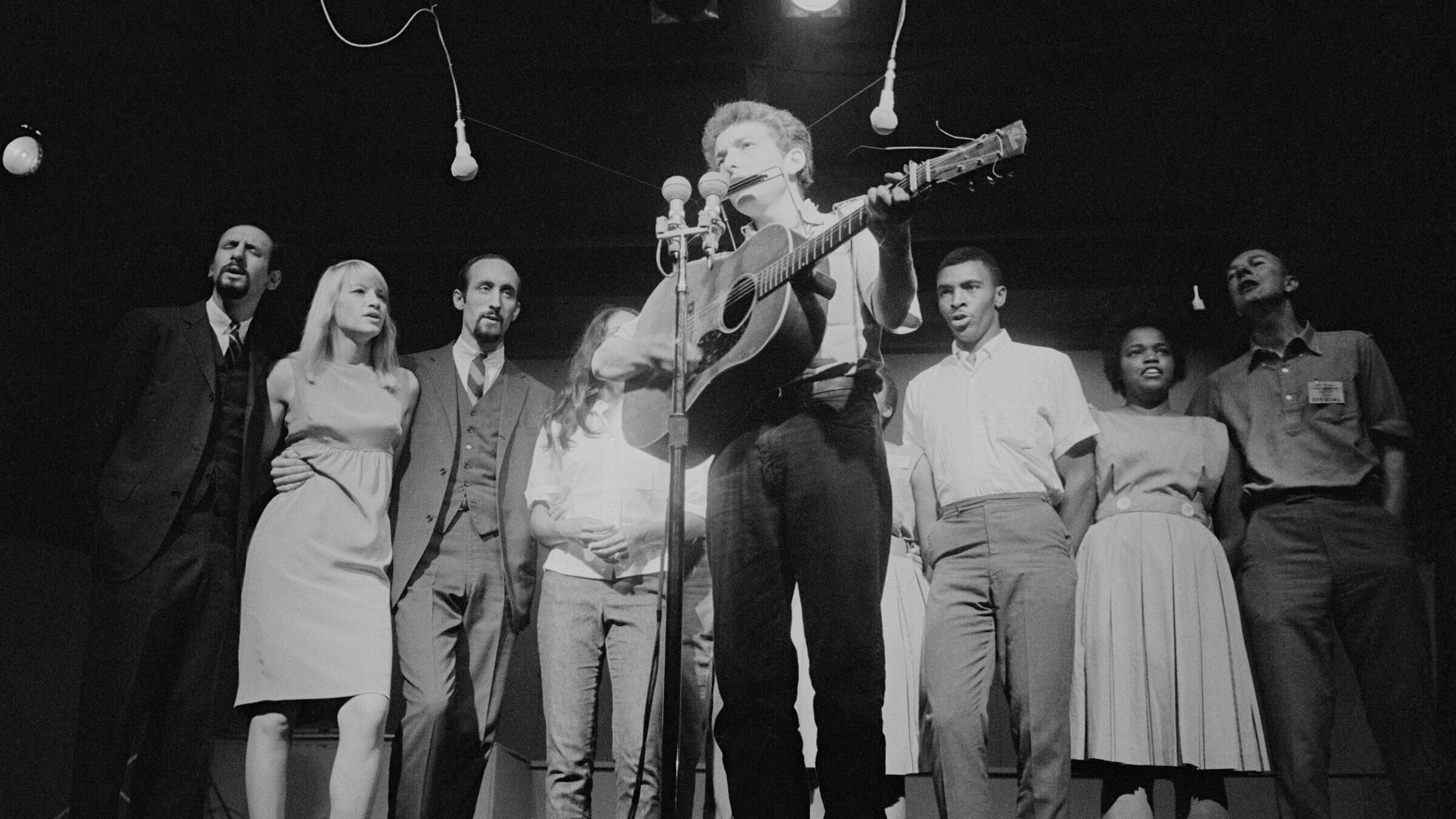 Bob Dylan  at the Newport Folk Festival, 1963. Among those behind him are, from left, Peter Yarrow, Mary Travers, Paul Stookey, Joan Baez (partially obscured), two unidentified people, Charles Neblett, Rutha Harris of the Freedom Singers, and Pete Seeger.