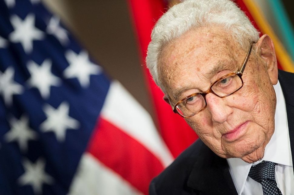 Former US Secretary of State Henry Kissinger, standing in front of an American flag.